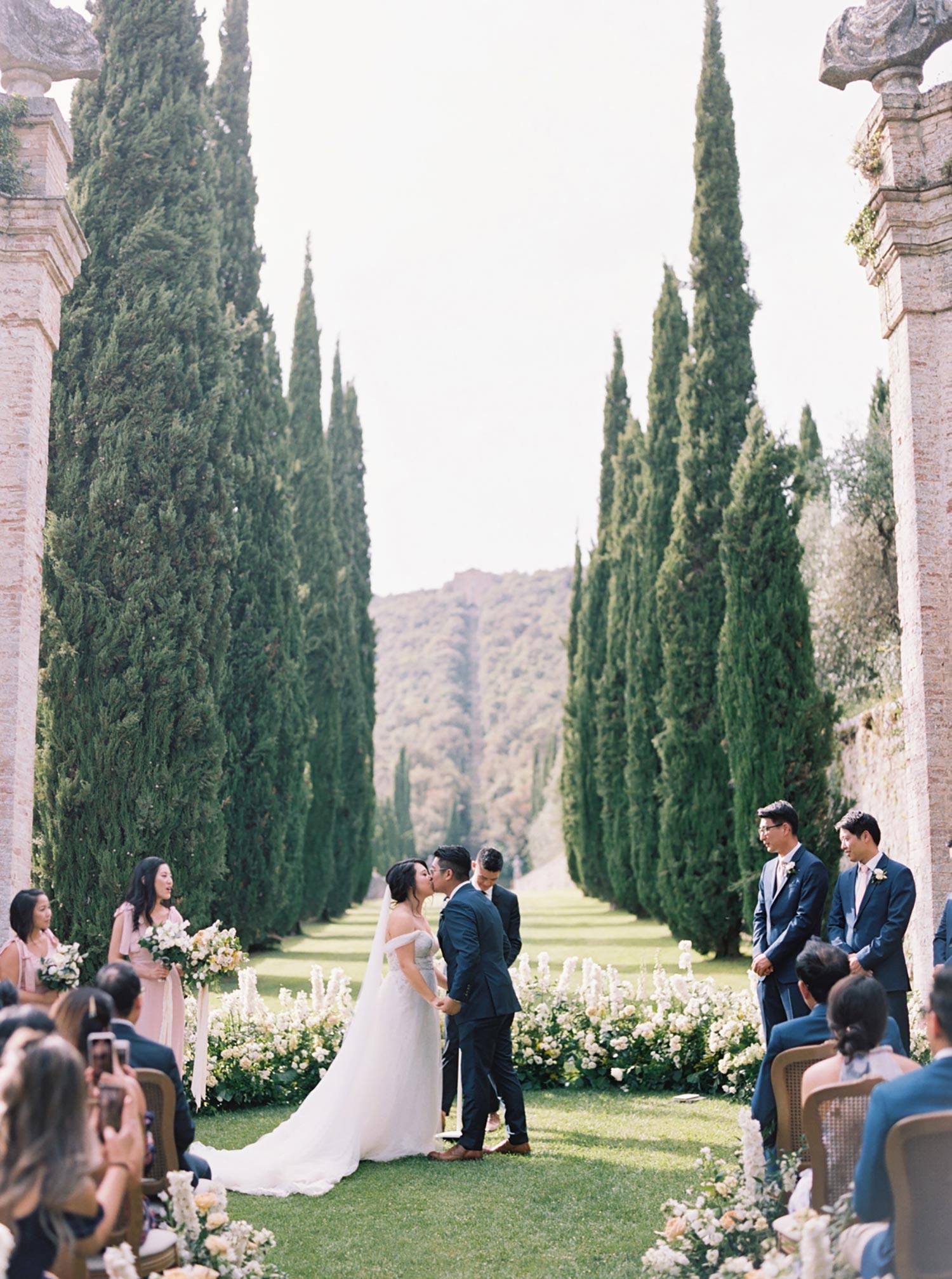 Italian villa wedding ceremony with cypress trees, cane back chairs and lush green and white flower aisle