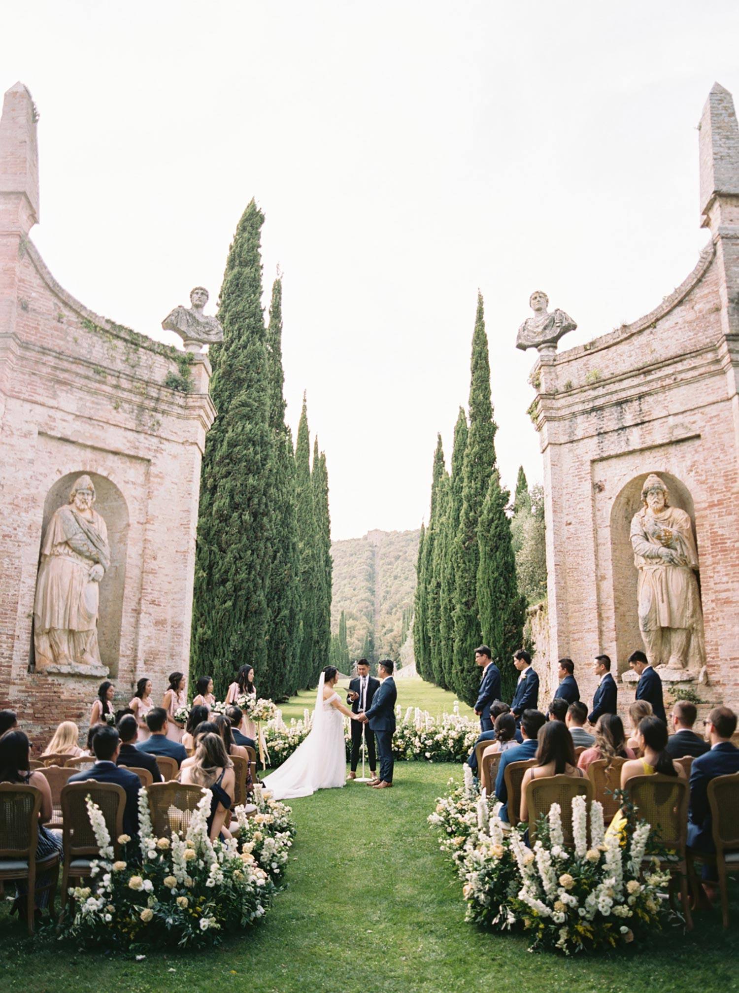 Italian villa wedding ceremony with cypress trees, cane back chairs and lush green and white flower aisle