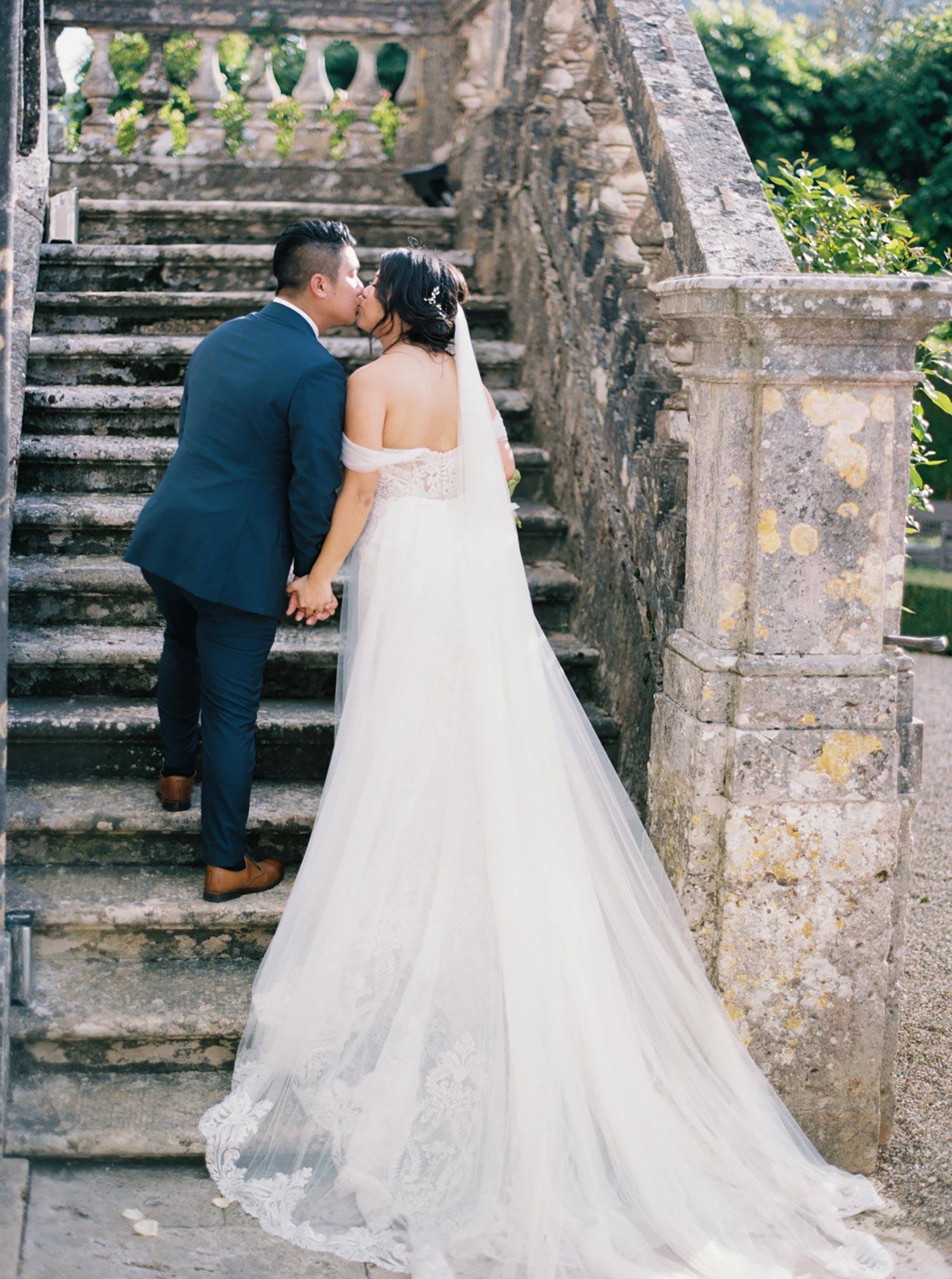 villa italienne historique avec vieil escalier en pierre et mariée dans une robe de mariée en tulle brodé à épaules dénudées embrassant le marié dans un costume de marié bleu marine