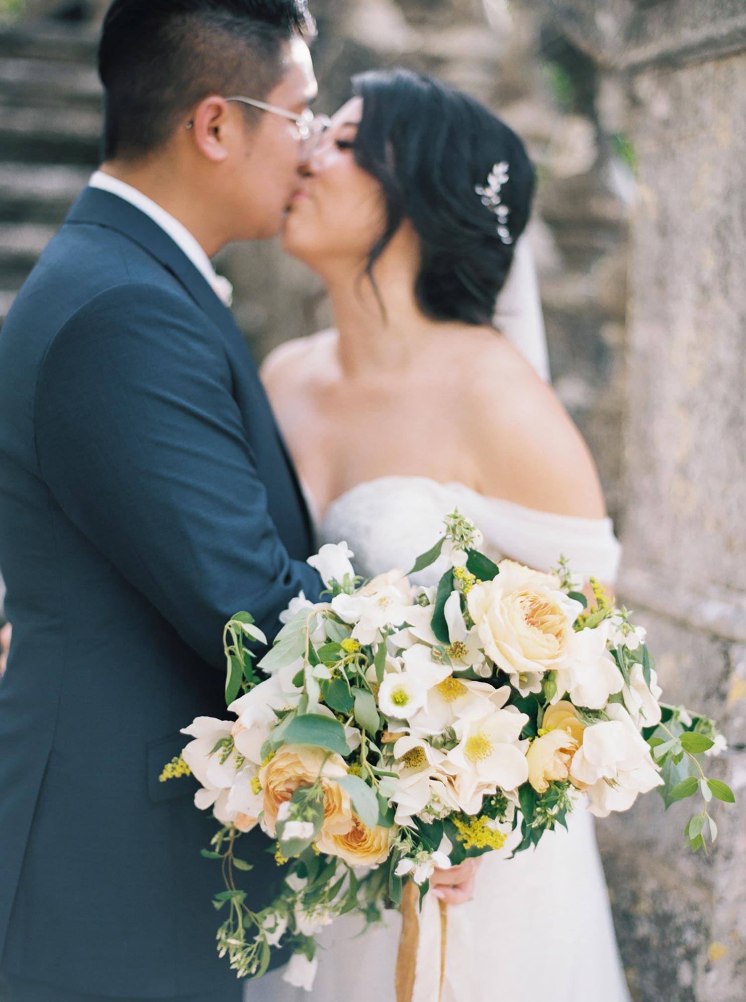 historic Italian villa with old stone staircase and bride in an off-the-shoulder embroidered tulle wedding dress kissing groom in a navy groom suit while she holds a yellow and white garden bridal bouquet