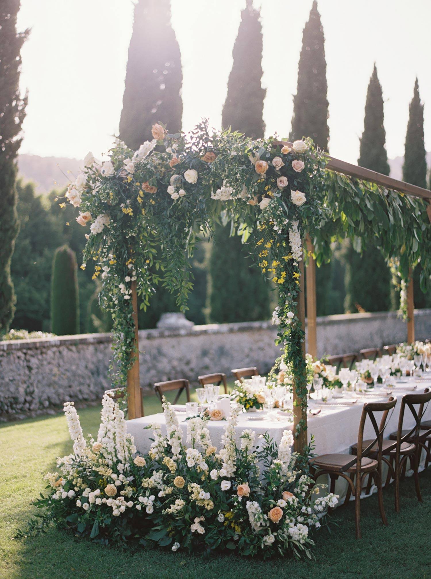 Dîner de mariage en plein air avec un décor de table de style familial, des compositions florales au sol luxuriant et une structure en bois suspendant de la verdure et des fleurs au-dessus de toute la table