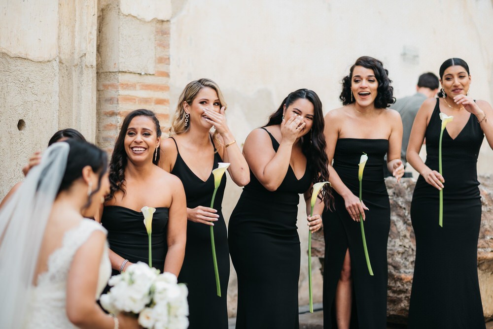 bridesmaids in floor length black gowns cheerfully react to brides dress