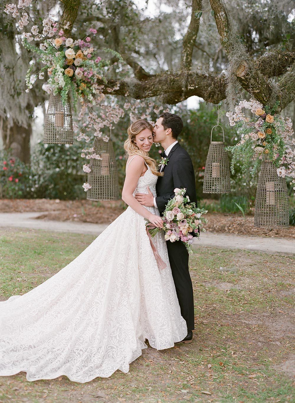 embroidered wedding dress with sweetheart neckline and black groom tux with live oak wedding backdrop and floral lanterns